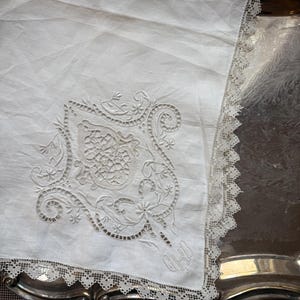 May include: A white linen handkerchief with intricate embroidered floral and scrollwork details. The handkerchief features a delicate lace trim along the edges and initials embroidered in the corner. The handkerchief is placed on a silver tray.