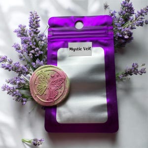 Może przedstawiać: Fioletowa torebka z napisem "Mystic Veil" na białej etykiecie, w towarzystwie okrągłego, dwukolorowego przedmiotu z motywem motyla. Gałązki lawendy otaczają scenę, tworząc spokojną, spa-podobną estetykę. Przedmiot z motylem jest zielony i różowy.