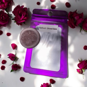 May include: A purple pouch with the text "Velvet Devotion" on a white label, accompanied by a circular, engraved pendant. The image is decorated with red rose buds and petals scattered on a white surface.