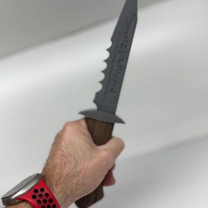 May include: A grey dagger with a serrated edge and a wooden handle is held. The blade has etched text. The person holding the dagger is wearing a red watch with a black perforated strap.
