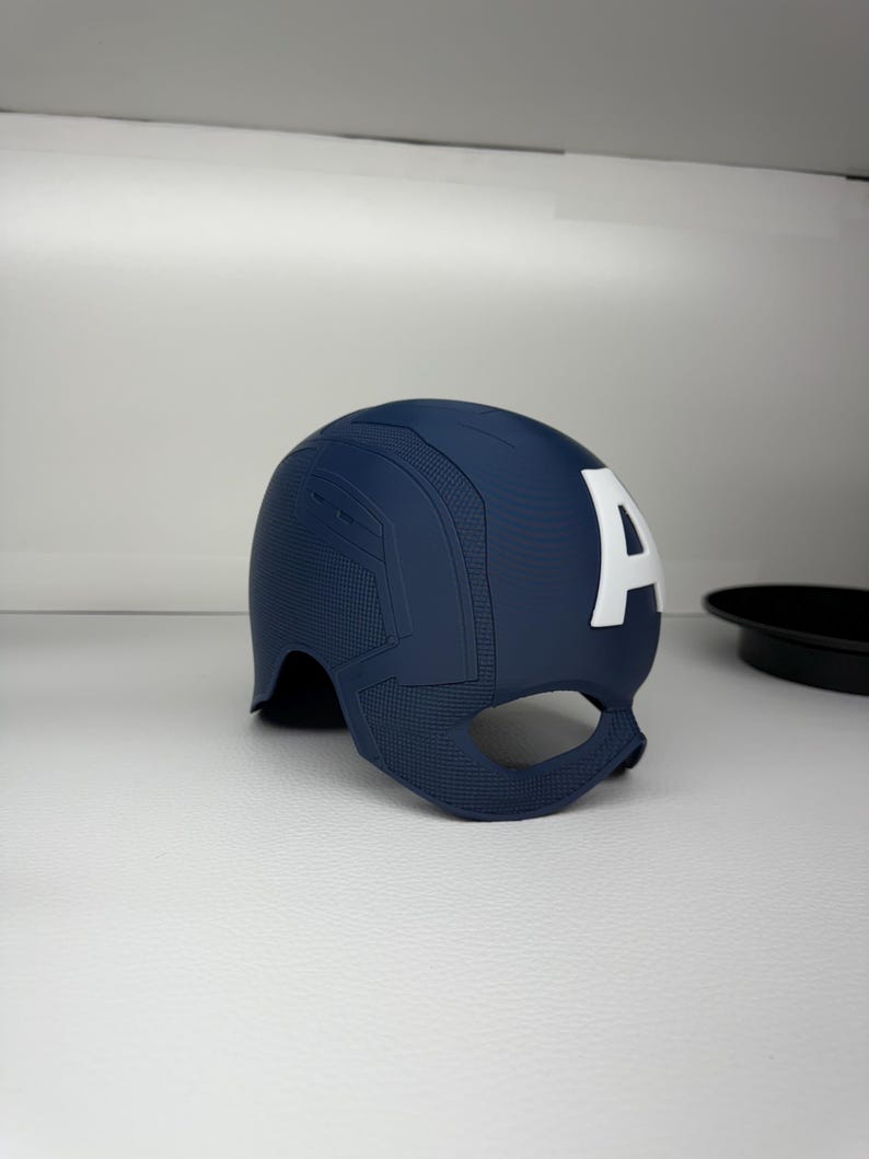 May include: A navy blue Captain America helmet with a white "A" on the side. The helmet has a textured surface and a chin guard. The superhero helmet is a replica of the one worn by Captain America.
