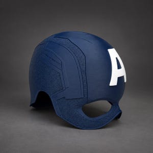 Può includere: Un casco blu scuro caratterizzato da una superficie testurizzata con linee di pannello modellate e una prominente lettera A bianca centrata sulla parte anteriore. Il casco presenta un'ampia apertura per occhi e naso e finitura opaca.