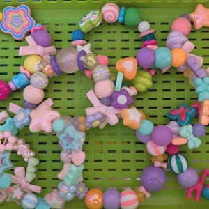 May include: A collection of colorful beaded bracelets in a green plastic basket. The bracelets feature various shapes, including stars, hearts, and bows, in pastel shades of pink, purple, blue, and yellow. The beads are of different sizes and textures.