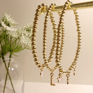 May include: Three gold beaded bracelets with charms. The charms include a small white shell, a gold letter L, and a gold star.