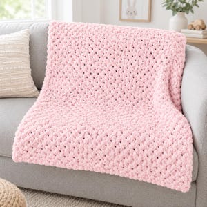Puede incluir: Una manta de punto grueso en color rosa claro, con una construcción de hilo texturizado y un patrón de bucles repetitivos. La manta está colocada sobre un sofá gris, mostrando su apariencia voluminosa y mullida con un diseño de puntada uniforme.