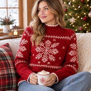 May include: A red long-sleeve knit sweater featuring a white snowflake pattern on the chest and sleeves, a high neckline, and ribbed trim. The sweater is paired with blue denim jeans and is held with a white mug featuring a red snowflake design.