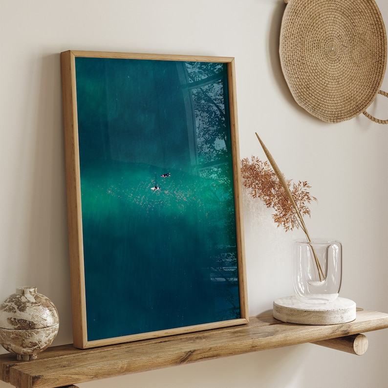 A peaceful home decor scene featuring a rustic wooden shelf with a textured vase and dried flora, beside a striking aerial photograph of two surfers adrift on a calm, sunlit ocean.