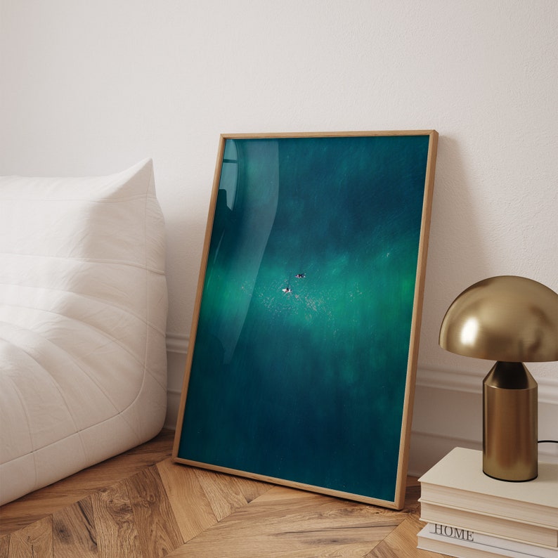 Warm bedroom ambiance with a leaning framed aerial shot of surfers on a teal ocean, next to a stylish metallic lamp and books on a wooden floor.