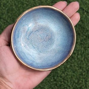 Puede incluir: Un pequeño cuenco redondo de cerámica con un esmalte degradado moteado en azul y blanco en la superficie interior. El cuenco tiene un borde liso de tono dorado metálico que rodea toda la circunferencia de la abertura circular.