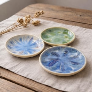 Könnte beinhalten: Drei kleine, runde Keramikschalen mit cremefarbenem Rand. Die Schalen haben ein blaues und grünes Aquarelldesign. Die Schalen sind auf einem neutralfarbenen Leinentuch angeordnet, das auf einem Holztisch liegt.