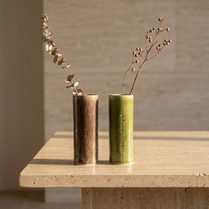 Peut inclure: Deux vases cylindriques, un marron et un vert, sont posés sur une surface en pierre claire. Chaque vase contient une tige de plante séchée. Le vase marron a un aspect texturé, le vase vert a un motif subtil.