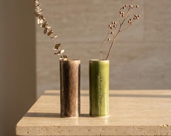 May include: Two cylindrical ceramic vases stand side by side. The left vase has a mottled brown finish, while the right vase is green with a vertical embossed tree pattern. Each vase holds a single dried botanical stem with small leaves or berries.