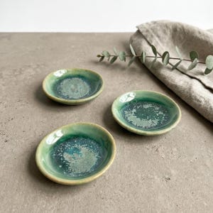 May include: Three small, round ceramic dishes featuring a reactive glaze with shades of deep forest green, teal, and light seafoam green. Each dish has an organic, slightly irregular rim and a smooth, glossy finish with a speckled texture.