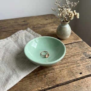 May include: A small, smooth, light mint green ceramic bowl with a circular gold-toned ring featuring a small clear stone resting inside. The bowl has a glossy finish and a simple, rounded shape, providing a clean surface for holding jewelry.