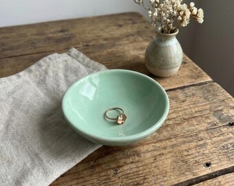May include: A small, smooth, light mint green ceramic bowl with a circular gold-toned ring featuring a small clear stone resting inside. The bowl has a glossy finish and a simple, rounded shape, providing a clean surface for holding jewelry.