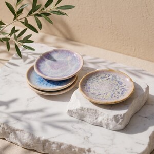 Peut inclure: Petites assiettes rondes en céramique avec un glaçage moucheté bleu, violet et crème. Les assiettes sont empilées sur une surface en marbre blanc. Un brin de feuilles vertes est visible en arrière-plan. Idéal pour les petites portions.