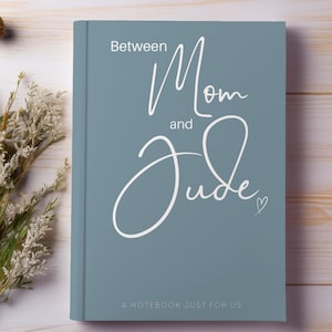 May include: A dusty blue hardcover notebook with white text that reads Between Mom and Jude, A NOTEBOOK JUST FOR US. The cover features a script font for the name Jude and a small heart icon. The notebook is positioned next to dried flowers.