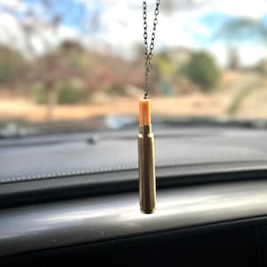 May include: A gold-colored bullet casing hangs from a silver chain, creating a rearview mirror charm. The casing has a light brown tip and is suspended against a blurred background of a blue sky with white clouds.