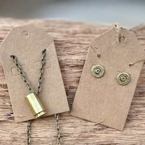 May include: A set of brass bullet jewelry, including a necklace with a bullet pendant and a pair of bullet earrings. The jewelry is displayed on two brown paper tags.