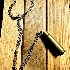 May include: A gold chain necklace with a brass bullet casing pendant. The necklace is lying on a wooden surface.