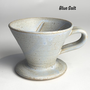 May include: A light blue ceramic pour-over coffee maker with a handle and a wide base. The words "Blue Salt" are written in black script at the top. The speckled surface gives it a rustic appearance.