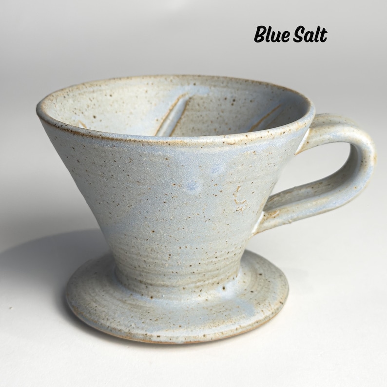May include: A light blue ceramic pour-over coffee maker with a handle and a wide base. The words "Blue Salt" are written in black script at the top. The speckled surface gives it a rustic appearance.