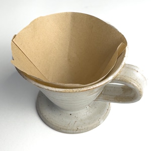 May include: A handmade ceramic pour-over coffee maker with a tan paper filter. The coffee maker is off-white with a speckled finish and a handle. The filter is folded and ready for use. Perfect for brewing coffee.
