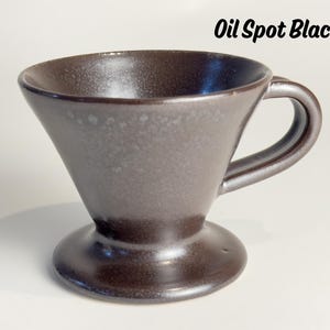 May include: A dark brown ceramic pour-over coffee maker with a handle. The cone-shaped brewer sits on a circular base. The text "Oil Spot Black" is in the upper right corner. The coffee maker has a speckled, textured surface.