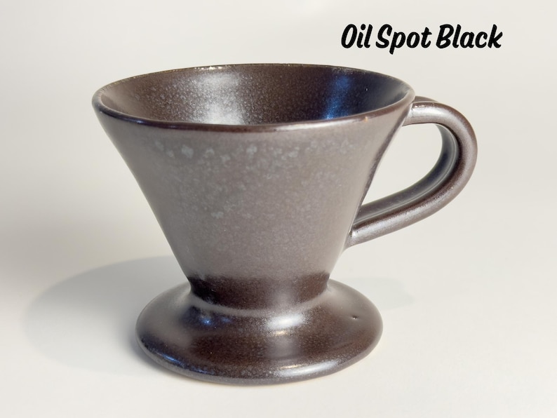 May include: A dark brown ceramic pour-over coffee maker with a handle. The cone-shaped brewer sits on a circular base. The text "Oil Spot Black" is in the upper right corner. The coffee maker has a speckled, textured surface.