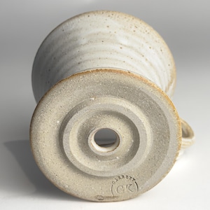 May include: A handmade ceramic mug with a light beige glaze and a textured surface. The mug has a rounded bowl shape and a small handle. The bottom of the mug has a circular base with a hole in the center and the text "Garrett (GK)".