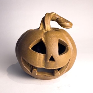 May include: A tan ceramic jack-o'-lantern with a curved stem, featuring triangular eyes, a small triangular nose, and a wide, toothy grin. The surface has a smooth, matte finish with subtle variations in color throughout the rounded body.