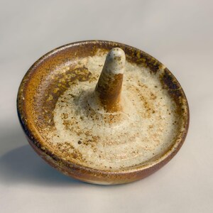 May include: A ceramic ring holder with a circular base and a central, upright post. The ring holder features a speckled, off-white interior and a brown, textured exterior. The piece is handmade and has a rustic, artisanal aesthetic.