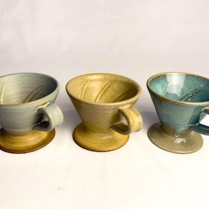 May include: Three ceramic coffee drippers with handles and circular bases. The left dripper is light blue, the centre is speckled tan, and the right is a glossy teal. Each features internal ridges and a wide, open top for brewing coffee.