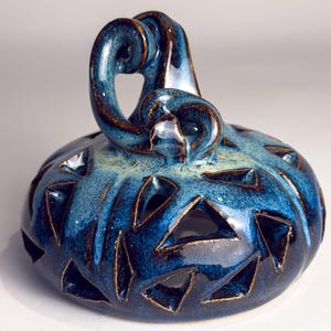 May include: A ceramic decorative piece with a blue and brown glaze. The piece has a rounded base with triangular cutouts and a curved handle. The glaze has a glossy finish, and the piece appears to be handmade.