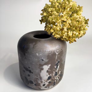 Könnte beinhalten: Eine dunkelgraue Keramikvase mit weißen und schwarzen Flecken, die einen Strauß getrockneter gelber Blumen hält.