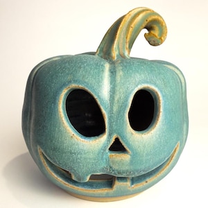 Op de afbeelding: Een blauwgroene keramische pompoen lantaarn met een glanzende afwerking en een gebogen steel. De pompoen heeft twee ovale ogen, een kleine driehoekige neus en een brede, uitgesneden mond met één tand zichtbaar aan de onderrand.