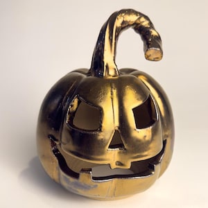 May include: A metallic gold ceramic pumpkin jack-o'-lantern with a curved stem. The pumpkin features cut-out triangular eyes, a small triangular nose, and a wide, jagged mouth opening. The surface has a reflective, glossy finish.
