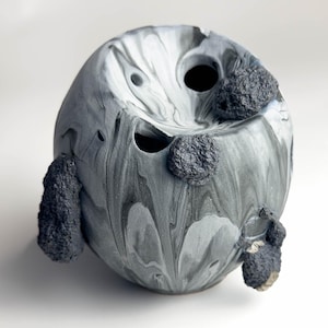 Puede incluir: Un jarrón esférico de cerámica con un patrón veteado gris y blanco. El jarrón presenta tres aberturas y varias protuberancias texturizadas de color gris oscuro. El diseño es abstracto y artístico.
