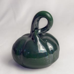 May include: A dark forest green ceramic pumpkin sculpture with a glossy, reflective finish. The pumpkin features a rounded, ribbed body and a prominent, curved, loop-shaped stem extending upward from the center of the top surface.