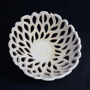 May include: A white ceramic bowl with a leaf-like cut-out design. The bowl has a scalloped edge and a textured surface. The bowl is centered on a black background. The bowl is likely handmade and suitable for decorative or functional use.