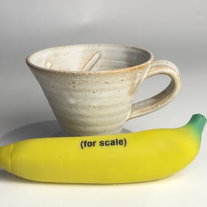 May include: A handmade ceramic coffee cup with a speckled, off-white glaze. The cup has a wide, angled bowl and a curved handle. A yellow banana with the text "(for scale)" is placed in front of the cup.