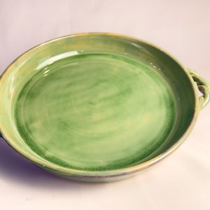 May include: A round ceramic serving dish with a glossy light green glaze and darker green accents. The dish features two twisted rope-style handles on opposite sides, with a smooth, shallow basin and a slightly raised rim around the edge.