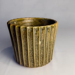 May include: A cylindrical ceramic planter featuring a deep olive green glaze with a textured, mottled finish. The exterior surface is defined by prominent vertical fluting that runs from the rim to the base, creating a rhythmic, ribbed pattern.