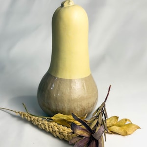 Könnte beinhalten: Eine zweifarbige Keramikvase mit gelbem Oberteil und beigefarbenem Sockel. Die vasenförmige Vase steht auf einer weißen Oberfläche, mit getrockneten Blumen und Blättern darum herum.