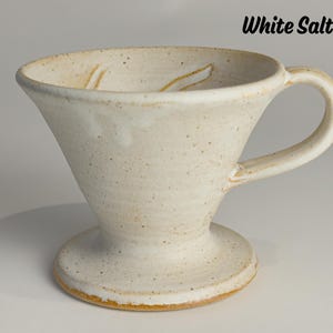 May include: Cream-colored ceramic pour-over coffee maker with a handle and a wide base. The cone-shaped body has a speckled texture and a slightly flared rim. The text "White Salt" is visible.