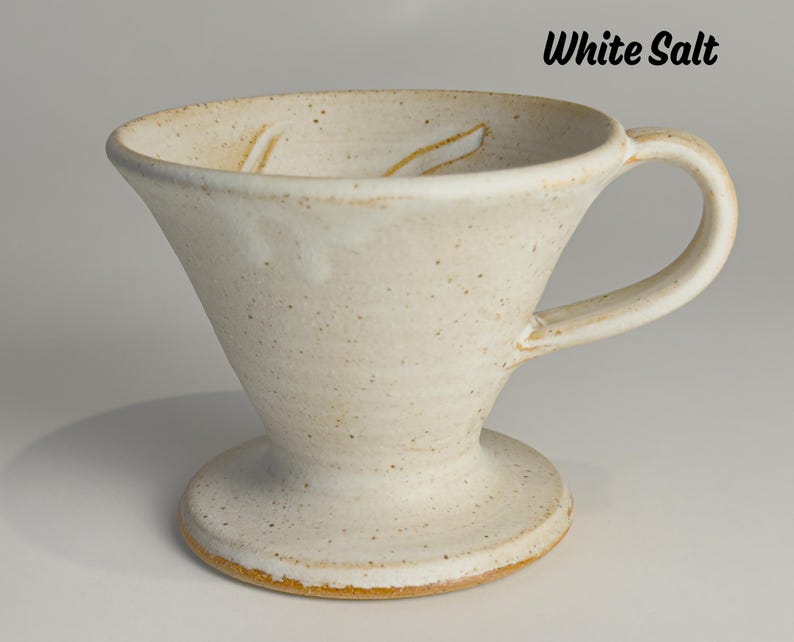 May include: Cream-colored ceramic pour-over coffee maker with a handle and a wide base. The cone-shaped body has a speckled texture and a slightly flared rim. The text "White Salt" is visible.