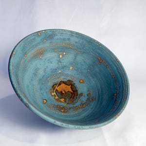 May include: A round ceramic bowl featuring a deep teal and blue glaze with a textured surface. The interior displays a mottled pattern of golden-brown speckles and a concentrated, dark amber-colored pool at the very center base.