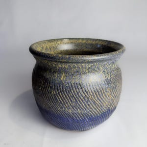 May include: A round ceramic pot featuring a textured surface with diagonal carved lines. The exterior displays a gradient color scheme, transitioning from a deep blue at the base to a mottled, earthy yellow and dark grey finish near the wide rim.