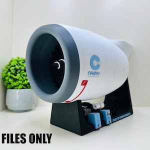 May include: A white and gray jet engine model mounted on a black stand. The engine features a dark gray intake, a black fan, and a blue logo with the text CADLY. The base includes small blue components and the text CADLY BY ADRIAN BARBOZA.