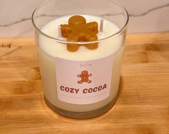 May include: A white candle in a clear glass jar with a gingerbread man topper. The label reads "Cozy Cocoa" with a small gingerbread man graphic.
